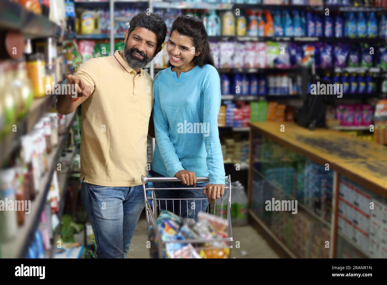 Felice coppia indiana che si diverte ad acquistare in un negozio di alimentari. Comprare della spesa per la casa al supermercato. Marito che punta verso lo scaffale. Foto Stock