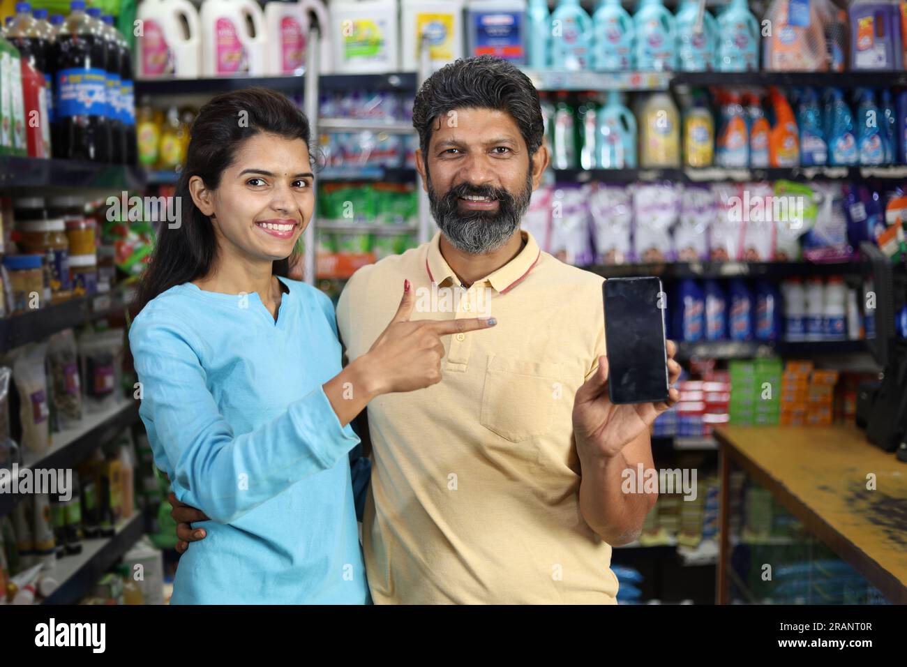 Felice coppia indiana che si diverte ad acquistare in un negozio di alimentari. Comprare della spesa per la casa al supermercato. Puntando verso lo schermo del cellulare Foto Stock