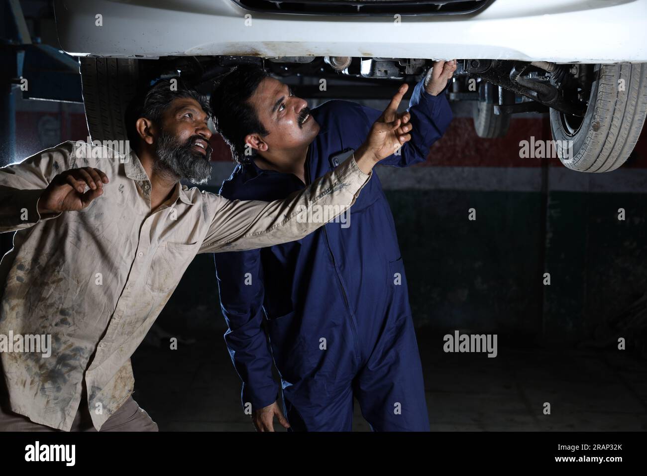 Meccanici in piedi e che lavorano insieme nella stazione di servizio. Specialisti che esaminano l'auto sollevata. Riparatori professionisti che parlano, fanno prestazioni. Foto Stock