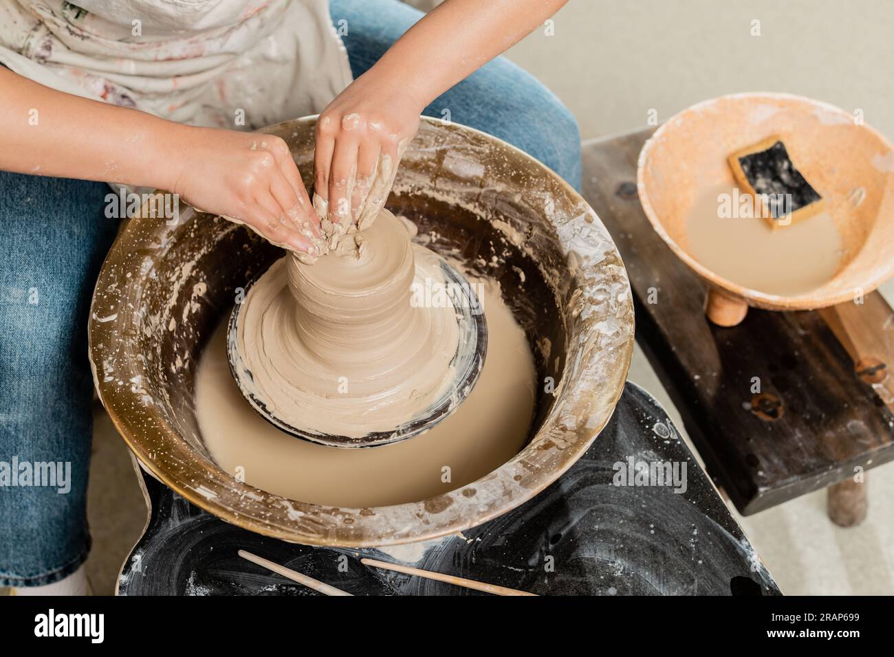 Vista ad angolo elevato di una giovane artigiana, che lavora in grembiule con l'argilla bagnata sulla ruota in ceramica e lavora vicino a una ciotola sfocata con acqua e spugna in una borchia in ceramica Foto Stock