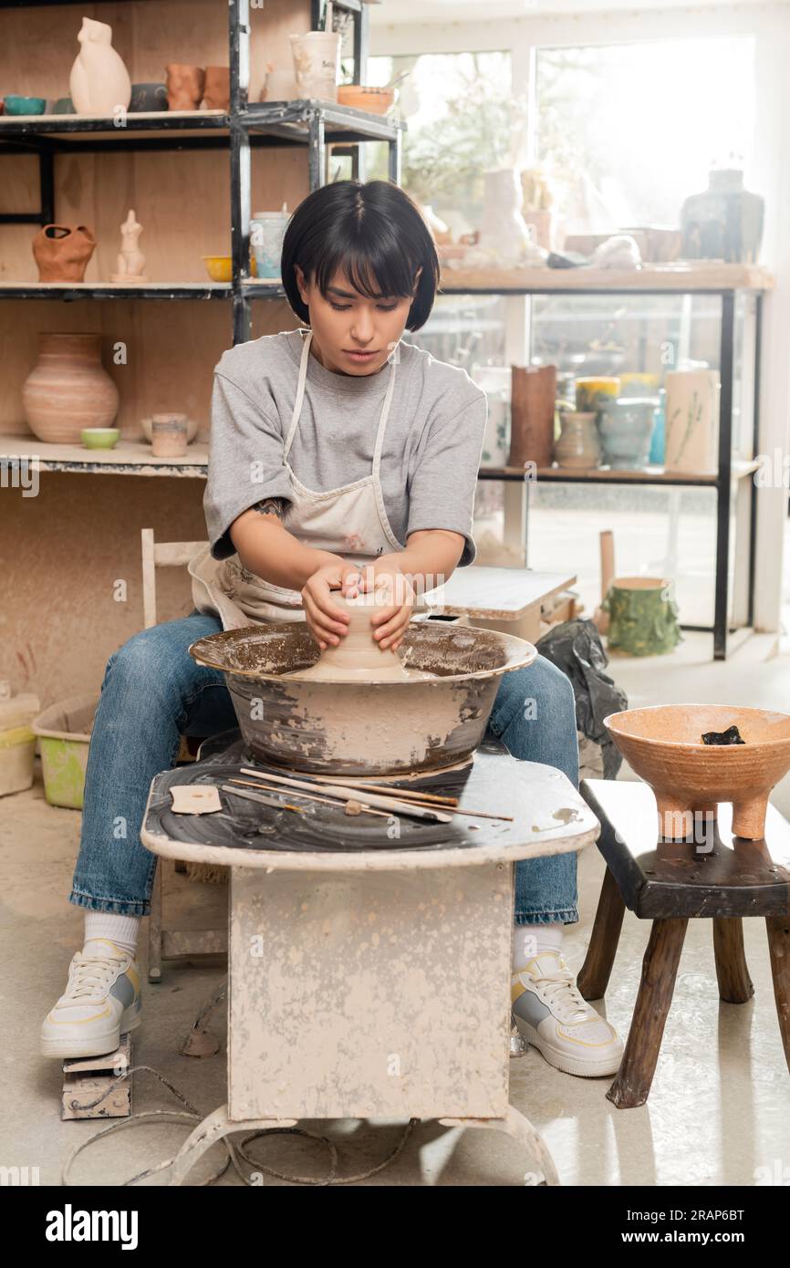 Giovane donna asiatica ceramista in grembiule che forma argilla bagnata su ruota in ceramica vicino agli attrezzi e ciotola con spugna mentre lavora nello studio di ceramica sullo sfondo, cl Foto Stock