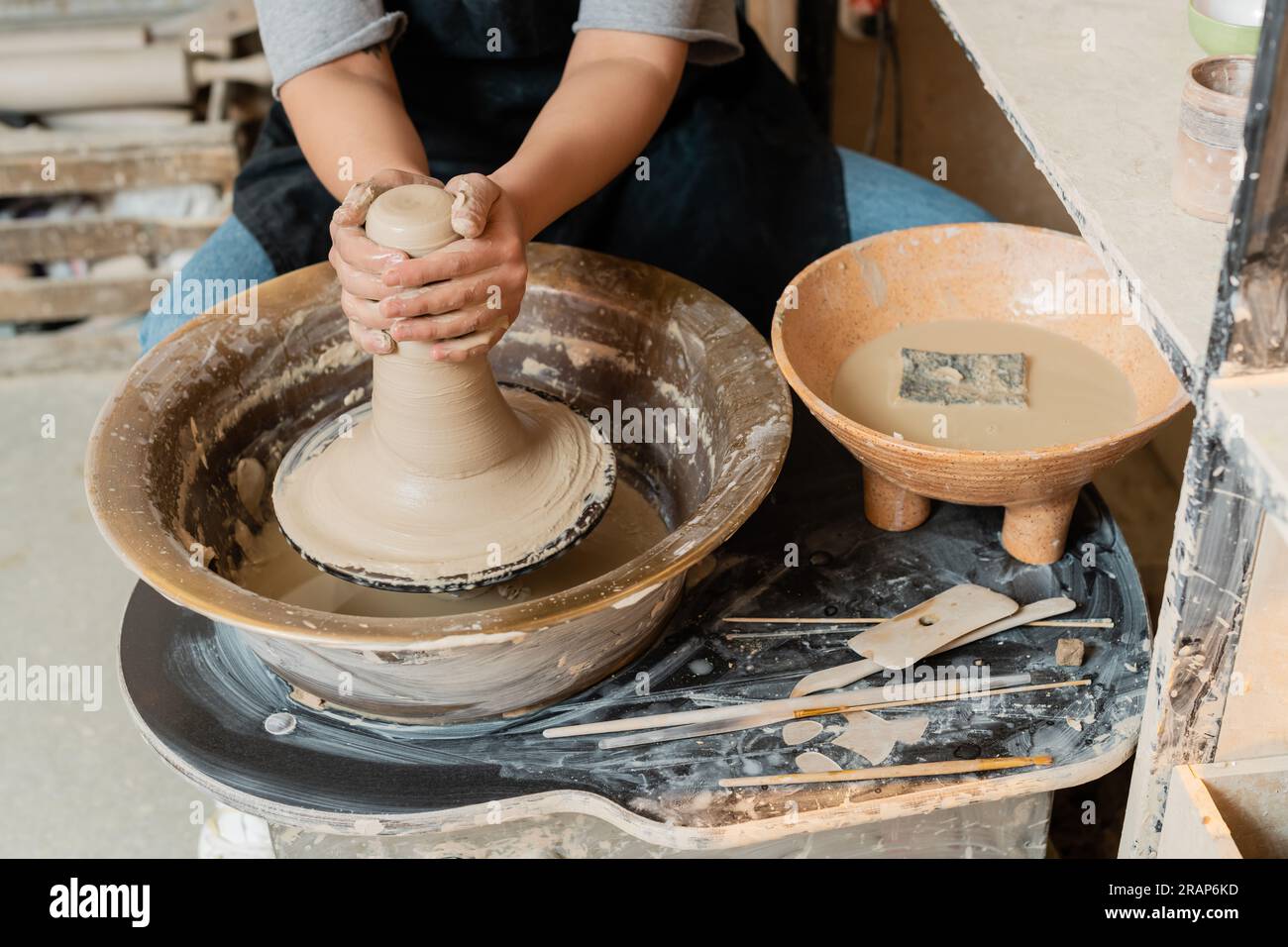 Vista ritagliata di una giovane artigiana in grembiule che forma argilla umida su ruota in ceramica vicino al recipiente con acqua, spugna e attrezzi sul tavolo in officina di ceramica, p Foto Stock