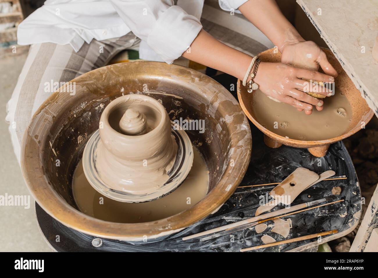 Vista dall'alto di una giovane artigiana in abbigliamento da lavoro che tiene la spugna vicino alla ciotola con acqua e argilla bagnata sulla ruota di ceramica mentre lavora in un laboratorio di ceramica, pott Foto Stock