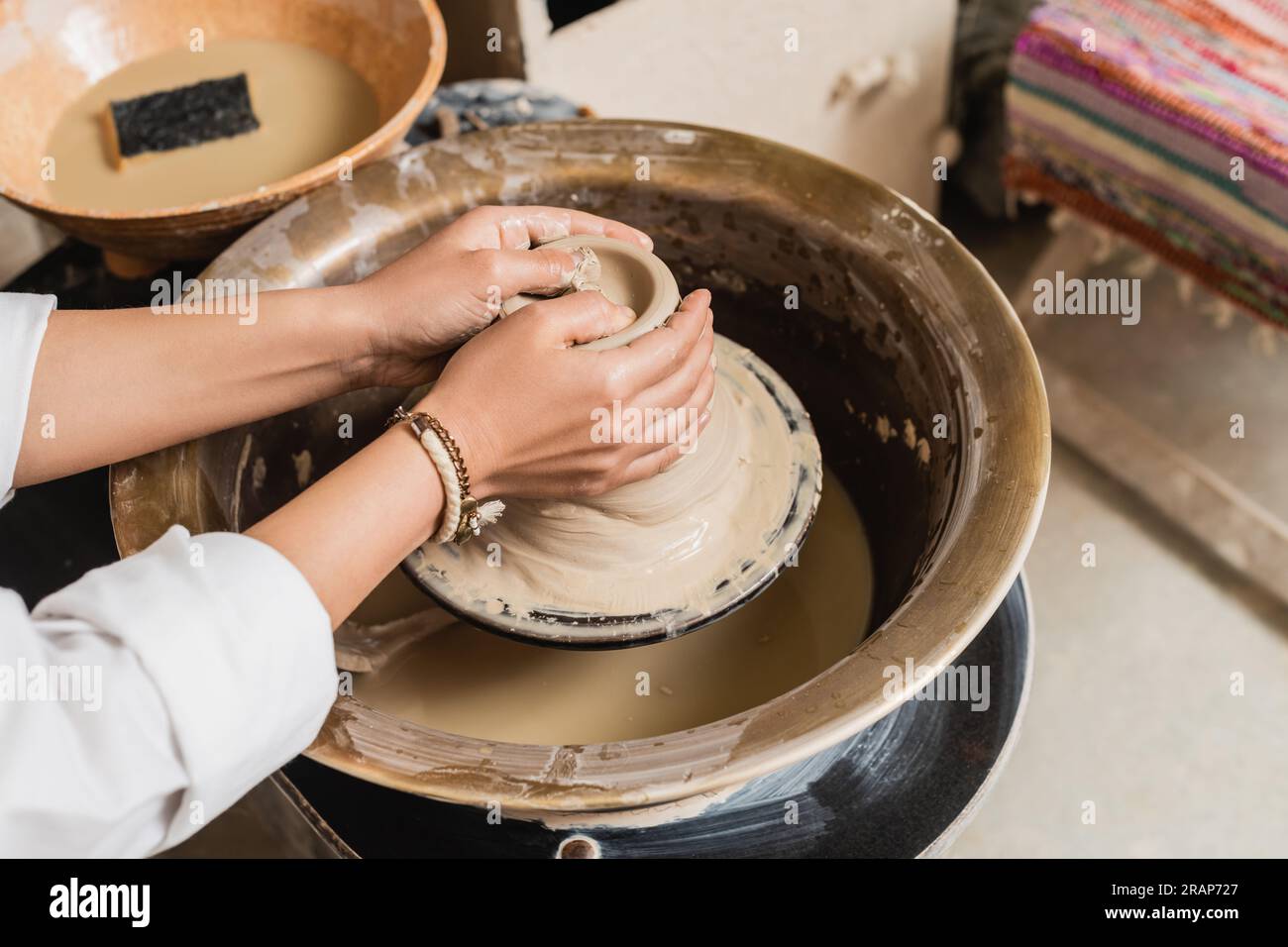 Vista ritagliata di una giovane artigiana che forma l'argilla bagnata su una ruota in ceramica vicino a un recipiente sfocato con acqua e spugna in un laboratorio di ceramica, un laboratorio di ceramica Foto Stock