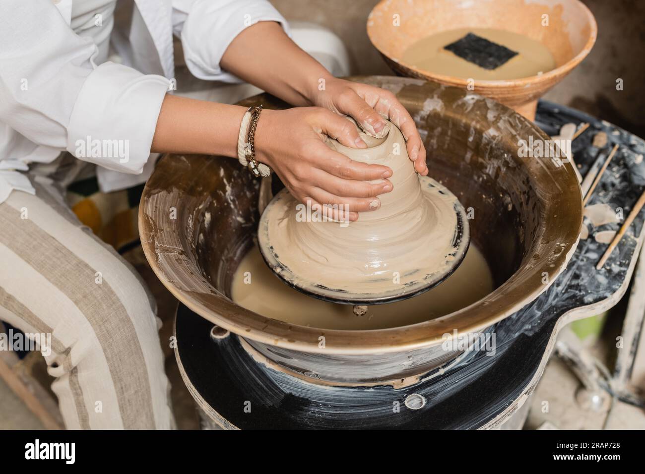Vista ritagliata di un giovane artigiano in abbigliamento da lavoro che forma l'argilla umida mentre lavora su una ruota in ceramica vicino a una ciotola sfocata con acqua e spugna sullo sfondo, in ceramica Foto Stock