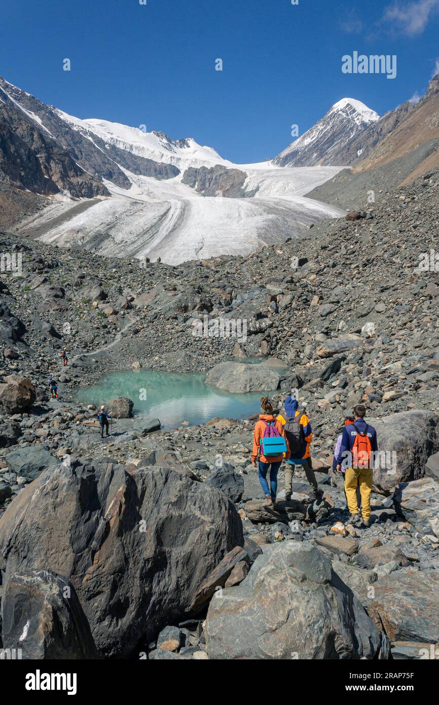Un gruppo di turisti di tre persone cammina lungo il sentiero verso un piccolo lago turchese e un ghiacciaio, le alte montagne di Altai, Aktru in estate Foto Stock