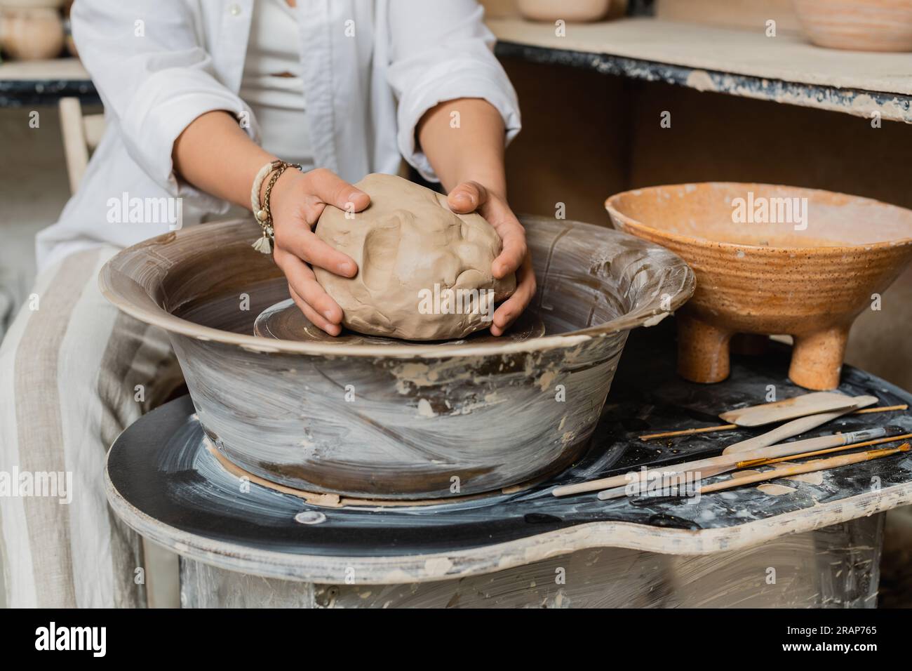 Vista ritagliata di una donna artigiana sfocata nell'abbigliamento da lavoro che mette l'argilla sulla ruota in ceramica vicino agli attrezzi e alla ciotola in un laboratorio di ceramica, scena di studio in ceramica con sk Foto Stock