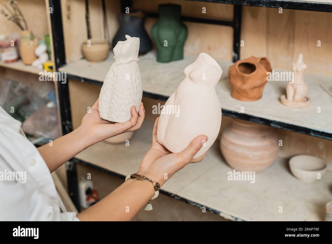Vista ritagliata di una giovane artigiana che indossa abiti da lavoro con sculture di argilla mentre si trova vicino a una griglia sfocata in un laboratorio di ceramica, scena di studio di ceramica wi Foto Stock