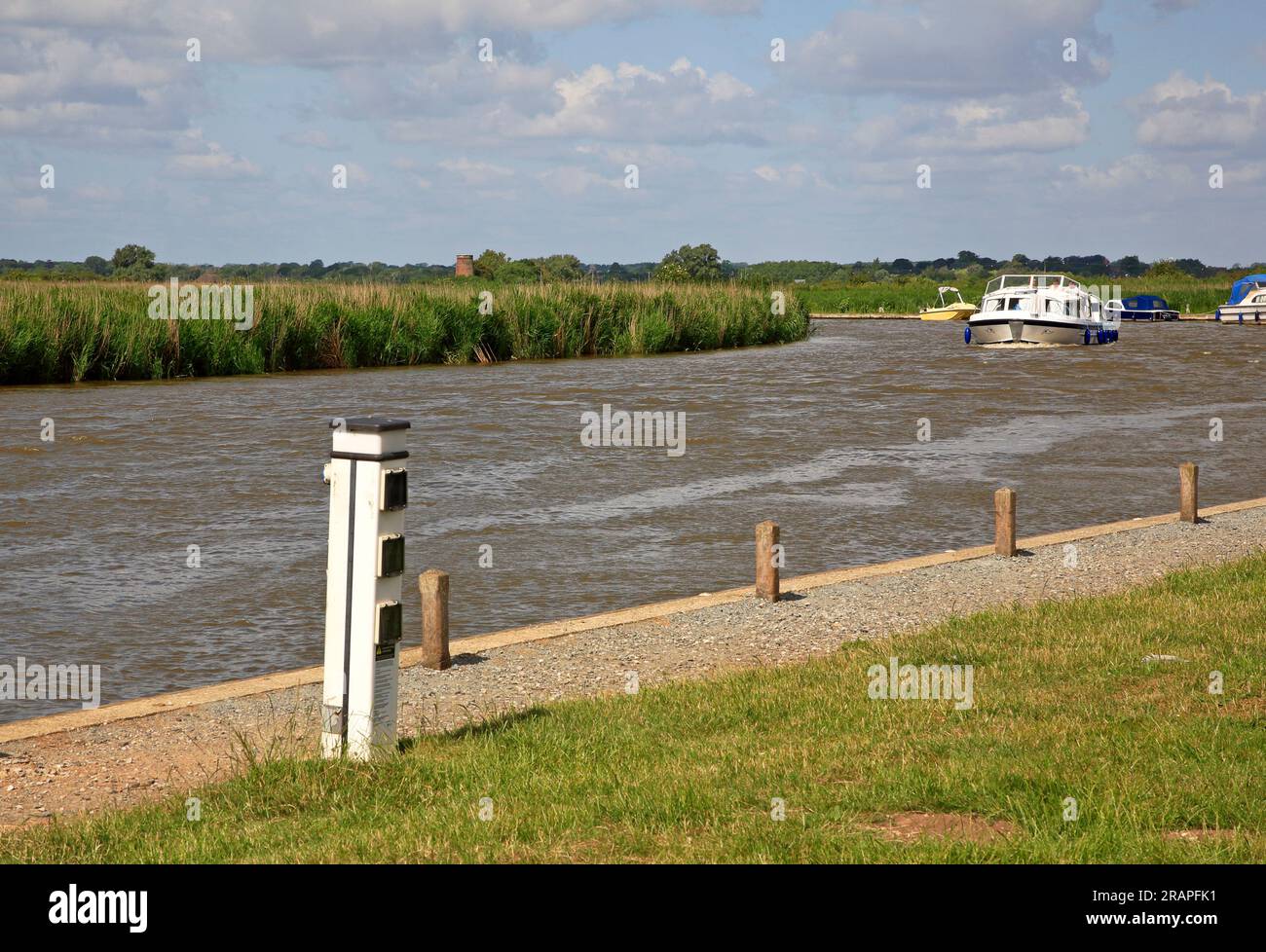 Una vista di uno Staithe con ormeggi e un incrociatore che si dirige sul fiume Bure sulle Norfolk Broads a Stokesby, Norfolk, Inghilterra, Regno Unito. Foto Stock