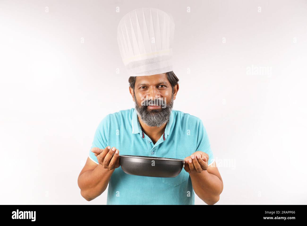 Ritratto di un cuoco indiano barbuto che indossa una t-shirt e un berretto da chef. Espressioni sorridenti su sfondo bianco. Tenere in mano la spatola e il cucchiaio per friggere Foto Stock