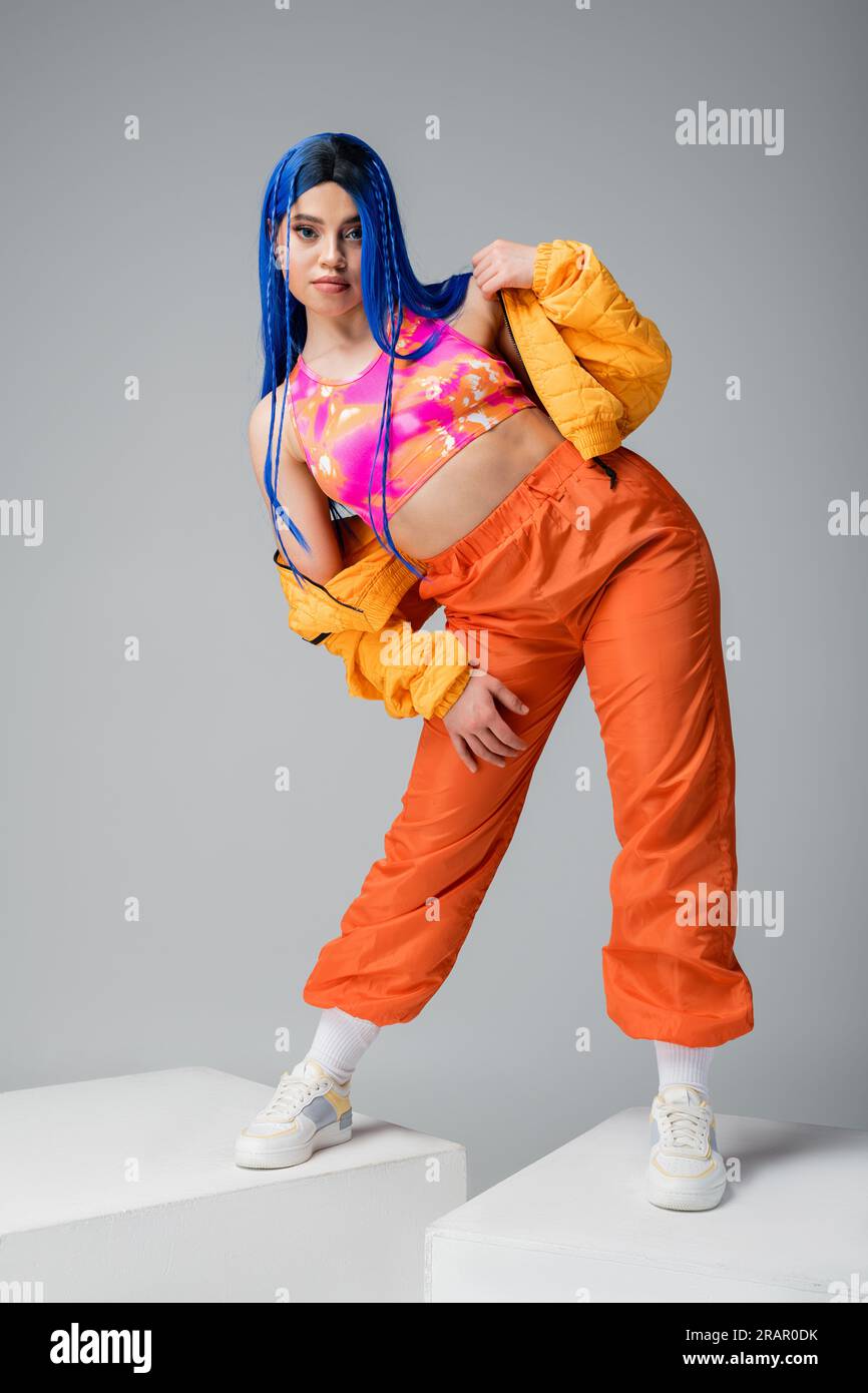 lunghezza intera, moda avanzata, donna con giacca a piumino regolabile con capelli blu e pantaloni arancioni su cubi bianchi sfondo grigio, colori vivaci, Foto Stock