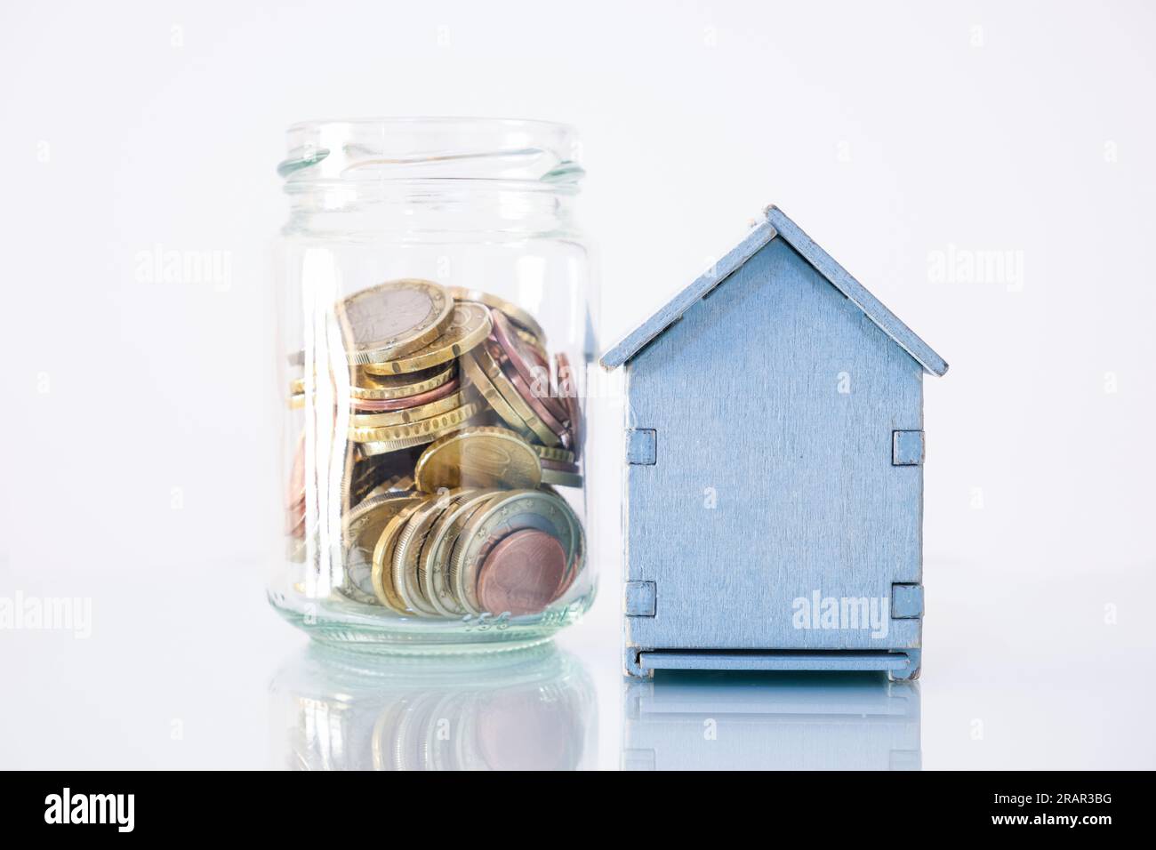 Una piccola casa di legno con accanto un contenitore di vetro in cui si accumulano le monete in euro, il concetto di risparmio, efficienza domestica. Foto Stock