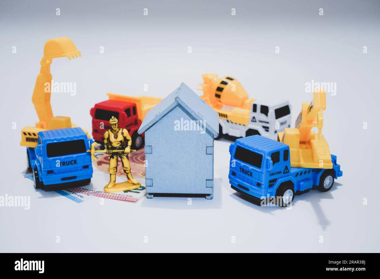 Una piccola casa che riposa sulle banconote in euro, nelle vicinanze ci sono veicoli e operai edili, concetto di costo per la ristrutturazione di una casa. Foto Stock