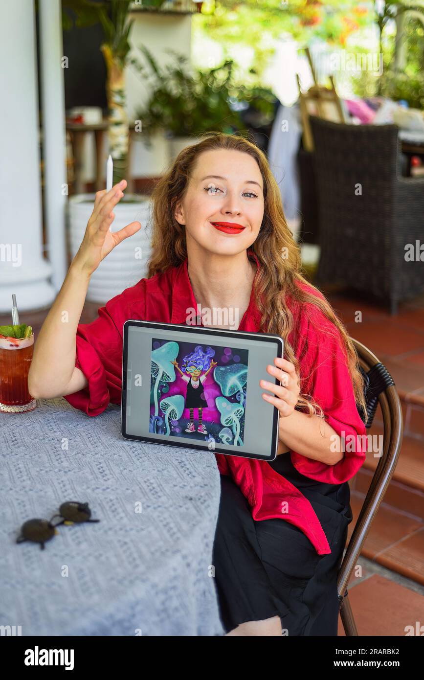 Bella artista e illustratrice bionda che mostra il suo disegno digitale, seduta accanto al tavolo con un tablet elettronico e uno stilo Foto Stock