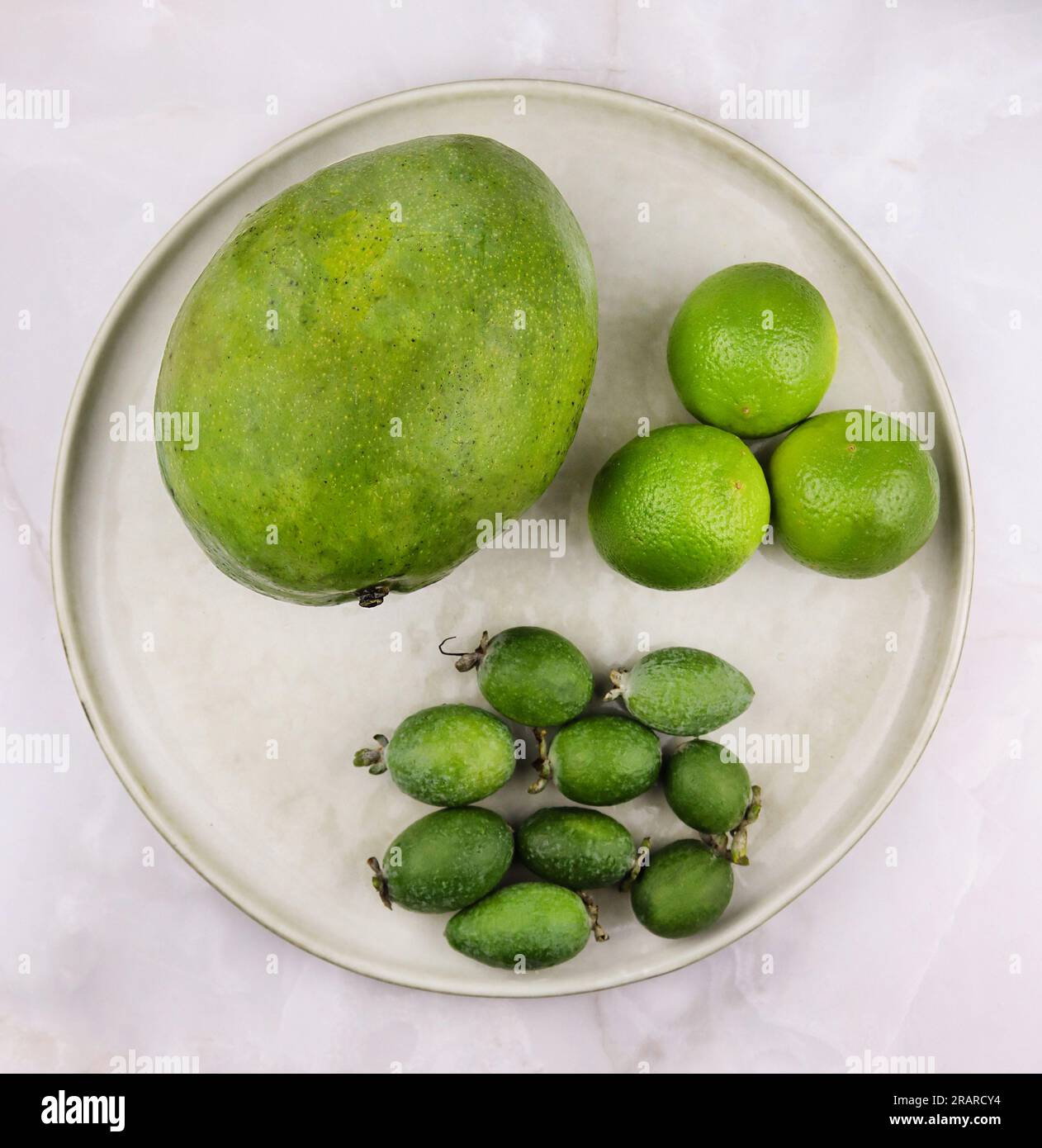 Frutto maturo di mango, bacche di feijoa intere e limi interi su un piatto isolato su fondo di marmo grigio. Trend del cibo verde. Supercibi esotici, cibo sano Foto Stock