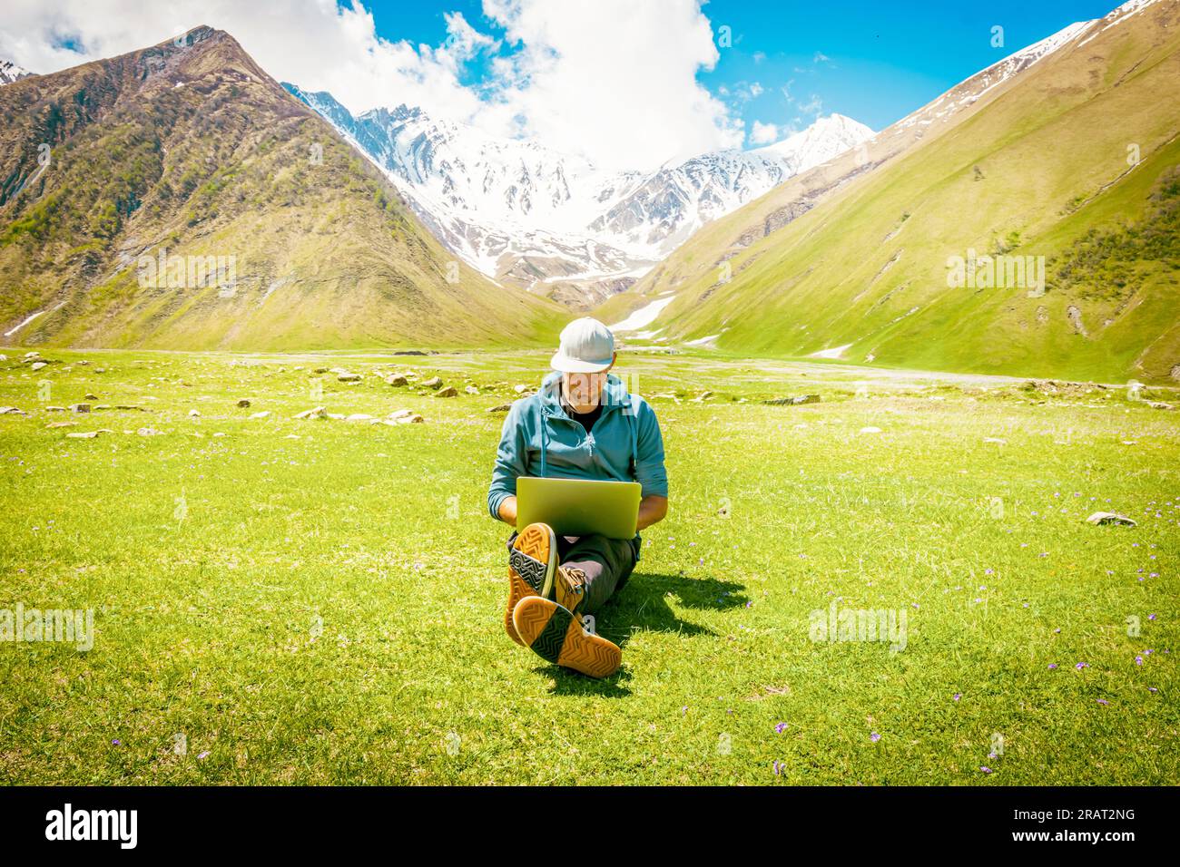 Uomo di mezza età con cappuccio bianco pieno siede con le gambe incrociate sull'erba con il laptop sulle ginocchia e digita con il sorriso in faccia. Lavoro a distanza Foto Stock