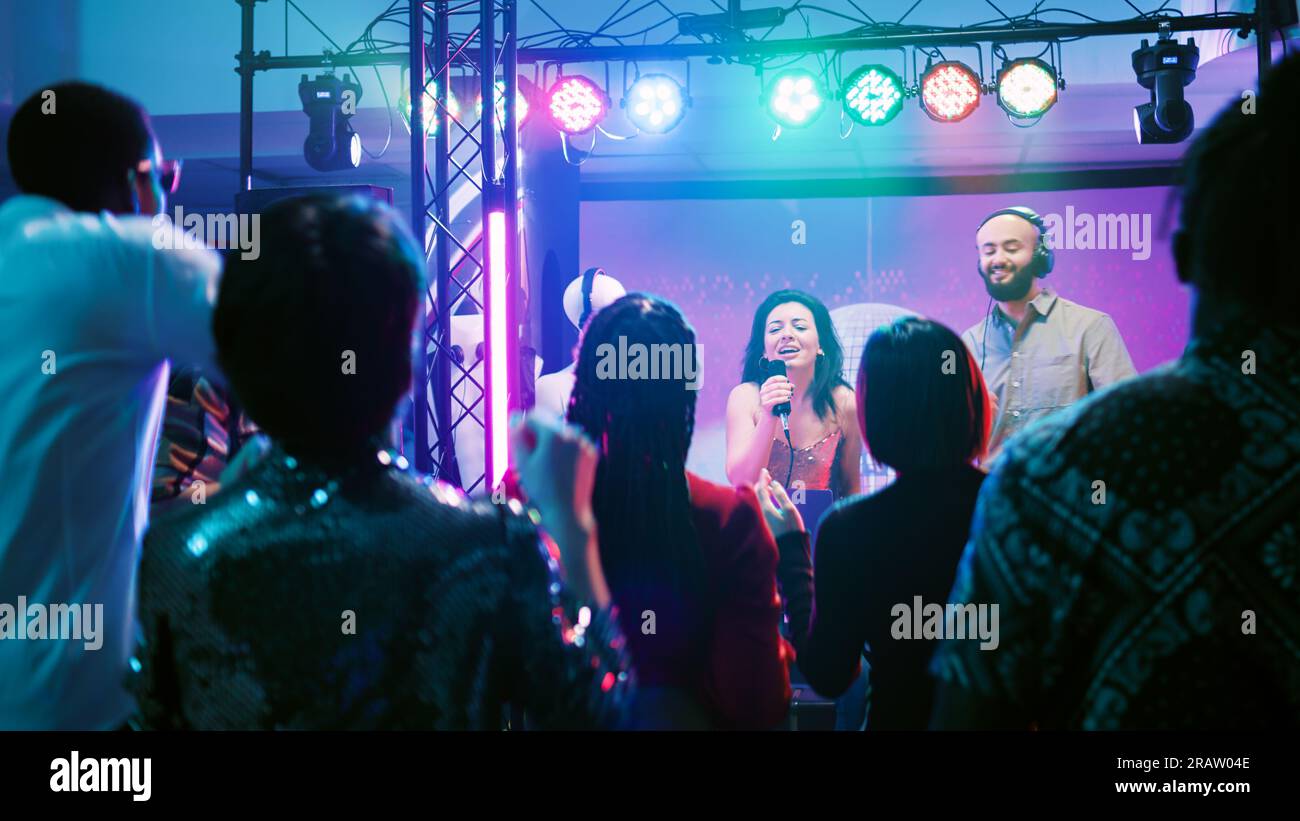 Ragazza sorridente che fa karaoke al club, canta canzoni al microfono e balla con DJ sul palco. Giovane che mescola musica per una folla di persone che si divertono e festeggiano, ballano. Scatto manuale. Foto Stock