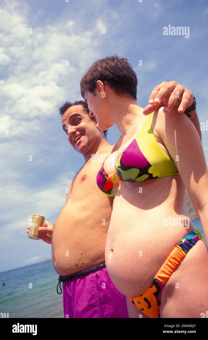 Donna incinta in bikini sulla spiaggia con un uomo che mostra la pancia Foto Stock