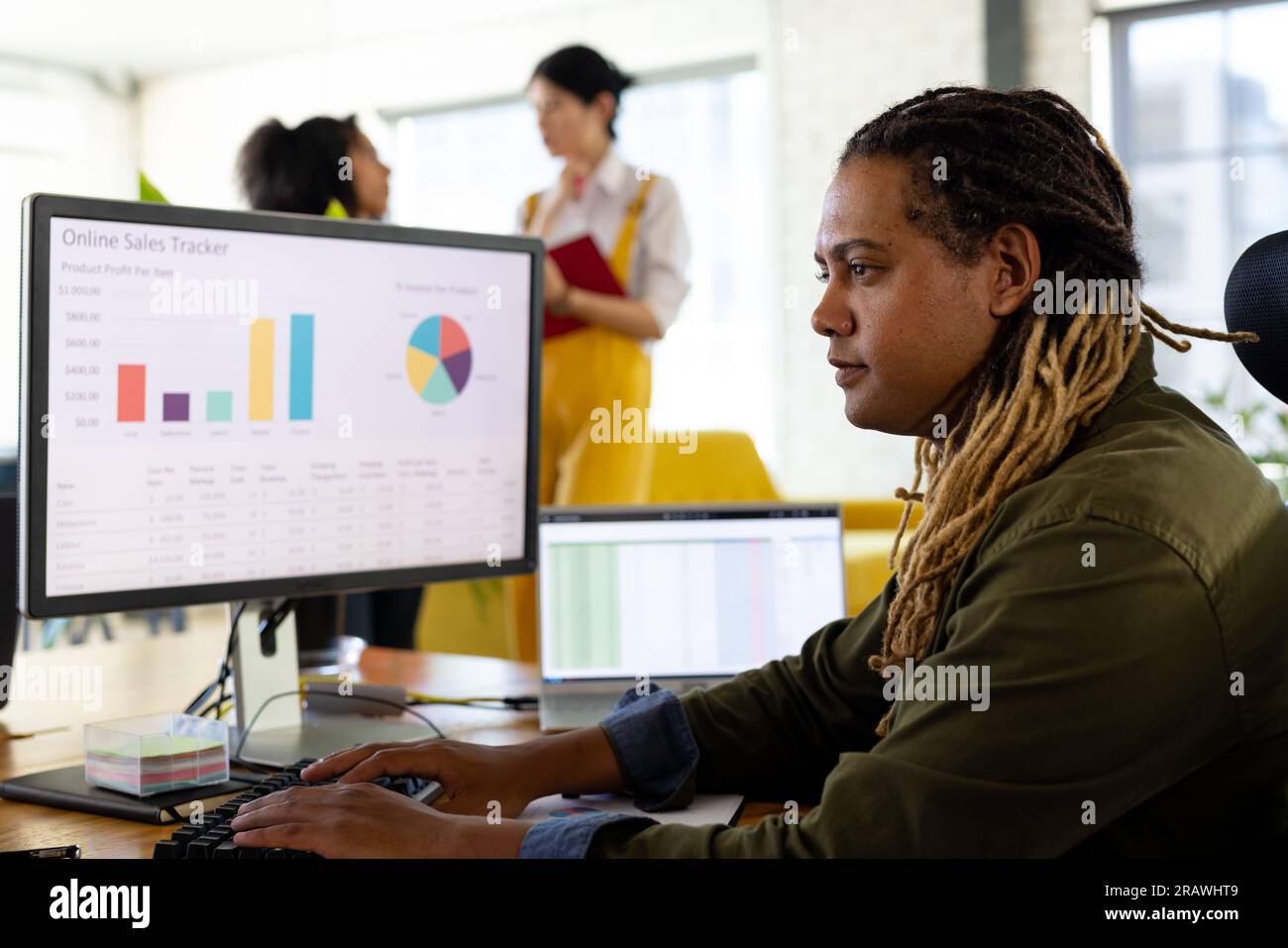 Uomo d'affari birazziale e informale con dreadlocks seduti alla scrivania che lavora al computer in ufficio. Lavoro, affari, comunicazione e ufficio informale, inalterato. Foto Stock