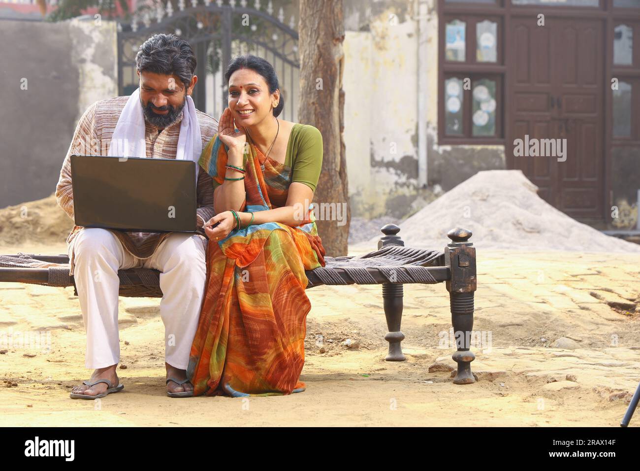 Marito e moglie indiane felici seduti insieme fuori dal cottage / casa / casa con un portatile in mano e navigando online. Foto Stock