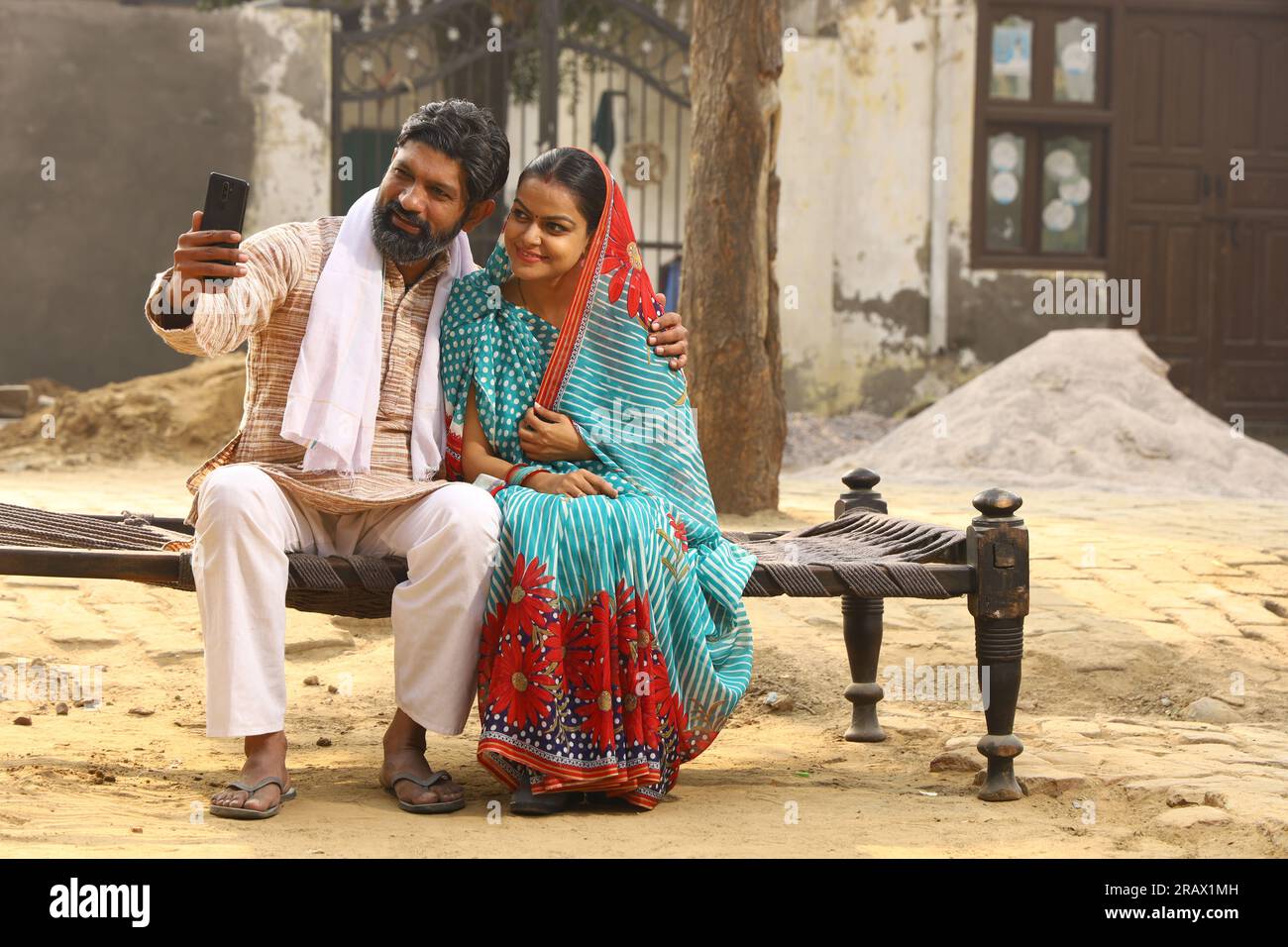 Una felice famiglia rurale indiana di marito e moglie seduti insieme fuori casa/casa/cottage, marito che mostra moglie al telefono cellulare. Foto Stock