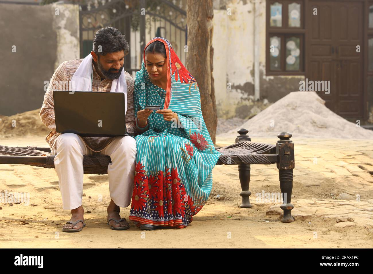Marito e moglie indiane felici seduti insieme fuori dal cottage / casa / casa con un portatile in mano e navigando online. Foto Stock