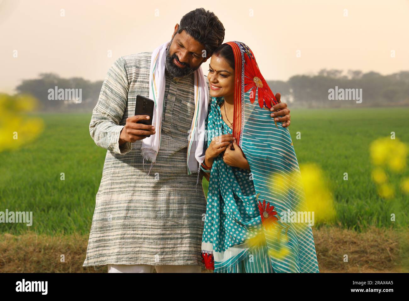 Una felice famiglia rurale indiana di marito e moglie seduti insieme fuori casa/casa/cottage, marito che mostra moglie al telefono cellulare. Foto Stock