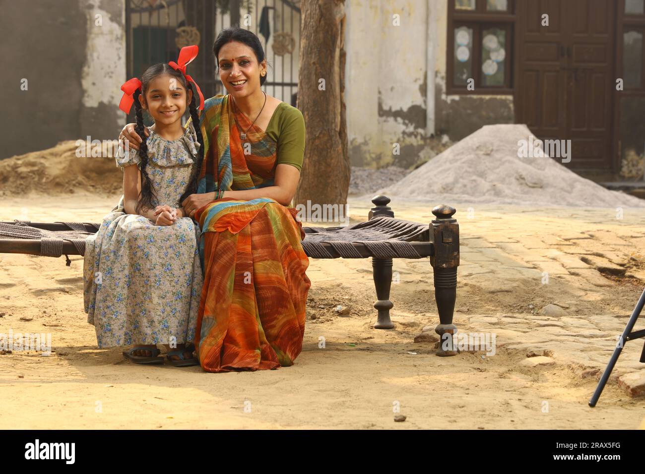 Mamma felice e figlie sedute insieme sorridendo sulla culla fuori casa, davanti al cortile. Madre in un bel sari. Cultura indiana. India rurale. Foto Stock
