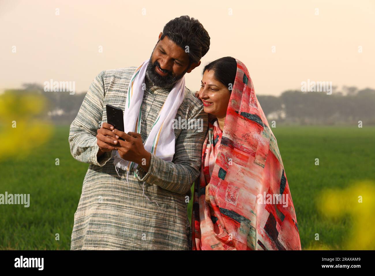 Una felice famiglia di abitanti di un villaggio rurale indiano in piedi in un marito di senape che mostra a sua moglie qualcosa sullo schermo del suo smartphone e ride Foto Stock