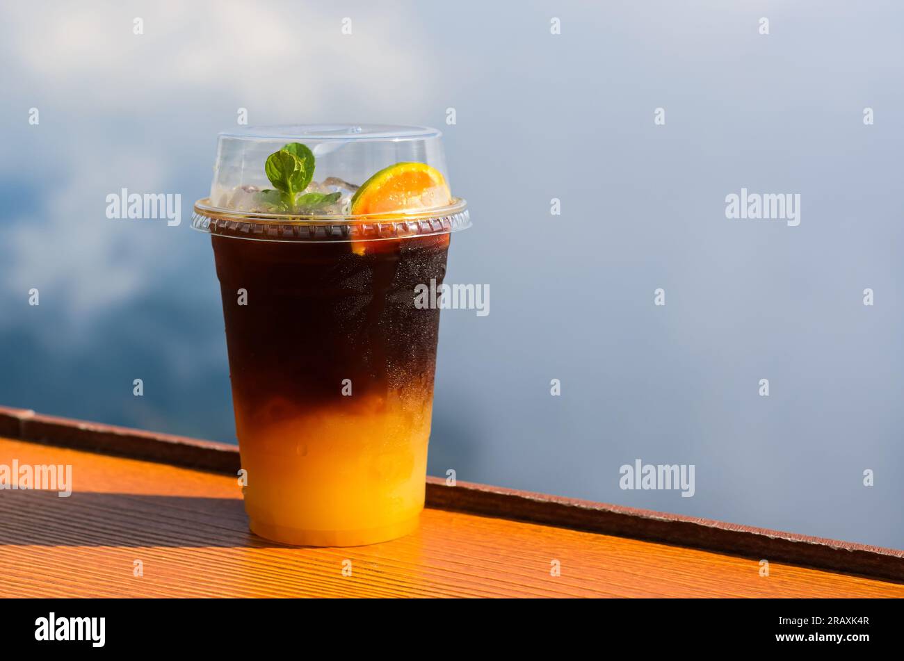 Un bicchiere di caffè ghiacciato si mescola con succo d'arancia messo sul tavolo con cielo e sfondo nuvoloso. Concetto di drink estivo. Foto Stock