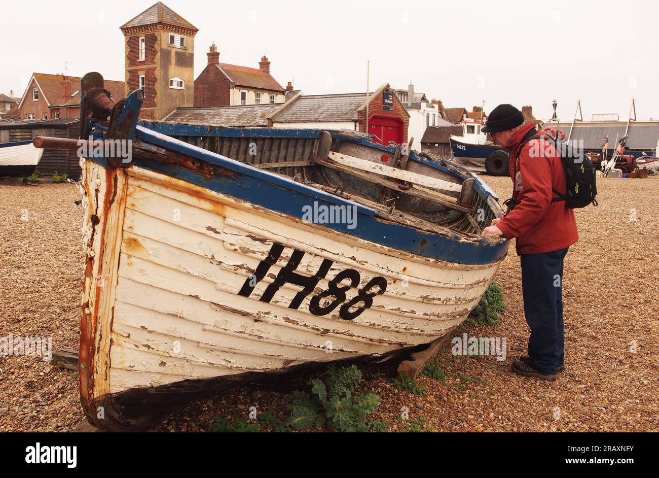 Un uomo più anziano di 60 anni, vestito con cappotto rosso, cappello di lana e con uno zaino che ispeziona una vecchia barca sulla spiaggia di ciottoli di Aldeburgh, Suffolk. REGNO UNITO Foto Stock