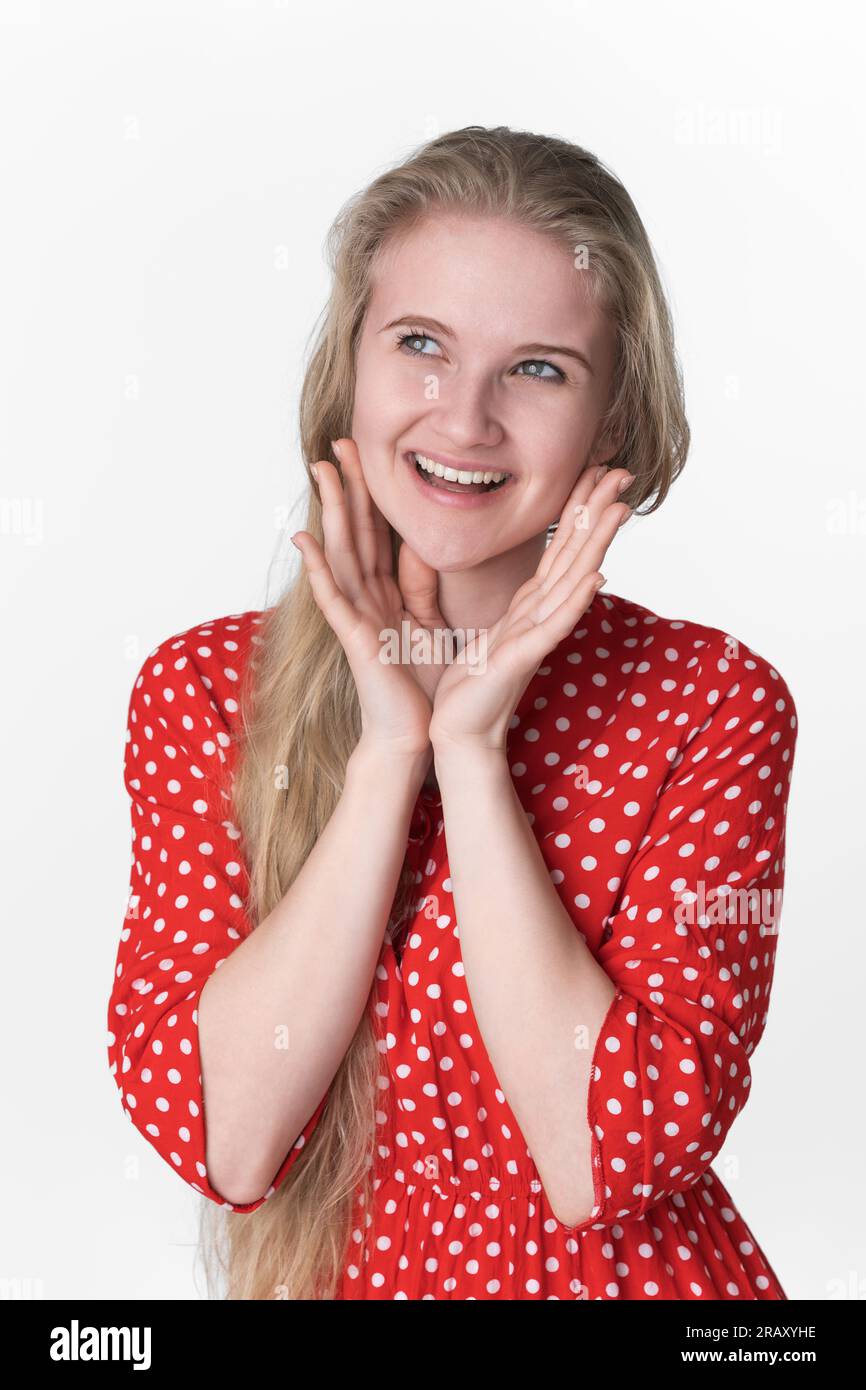 Ritratto di una giovane donna bionda adulta con emozioni positive sul viso, con palmo piegato sotto il mento, guardando lontano. Giocoso modello di 21 anni in abito rosso Foto Stock