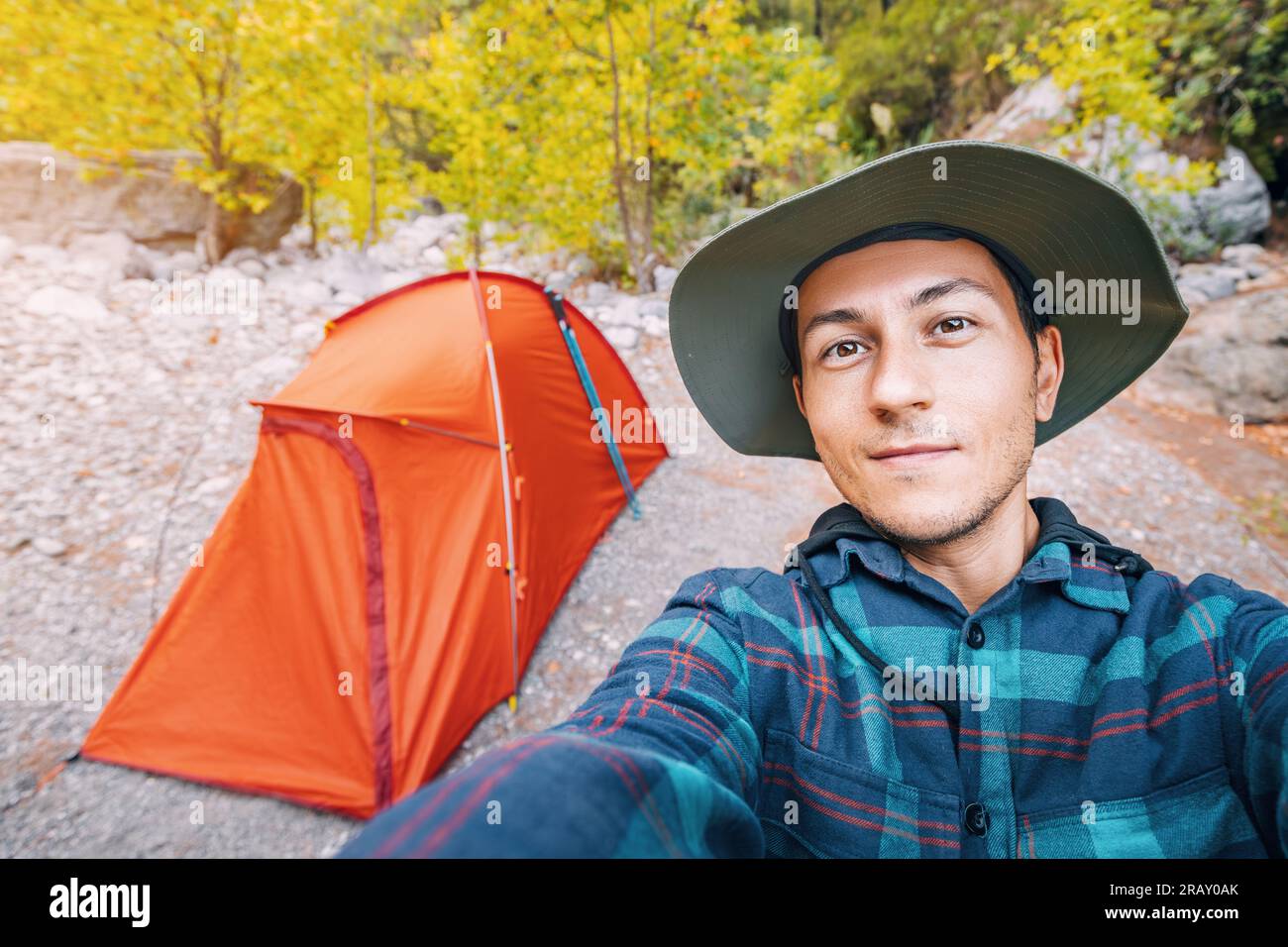 Uomo escursionista felice che indossa un cappello che scatta foto selfie contro una tenda rossa nel bosco. Attrezzatura per passeggiate e trekking da solo Foto Stock