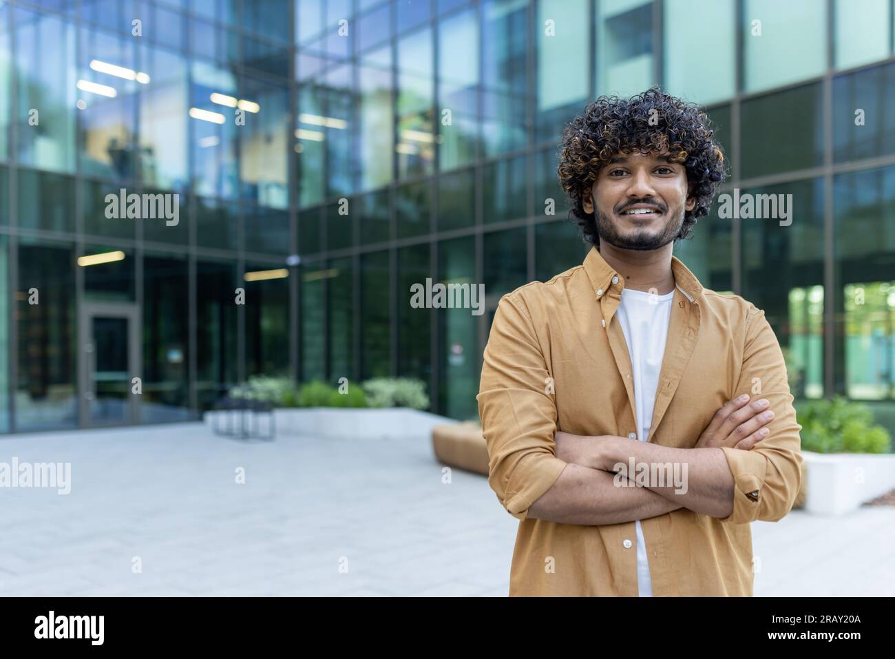 Ritratto di uno studente indiano giovane sicuro di sé, bello e intelligente che si trova all'esterno del campus con le braccia incrociate sul petto e sorridente davanti alla fotocamera. Foto Stock