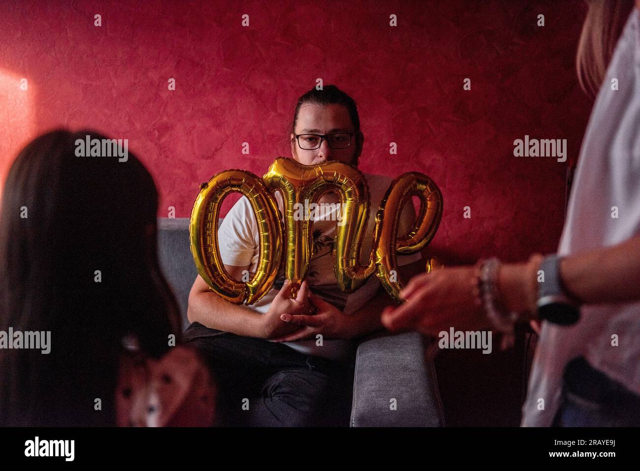 Il giovane uomo della diversità con gli occhiali festeggia tristemente il suo compleanno. Millennial che si aggira, sbircia fuori dal palloncino in oro laminato che ne indica UNO. Appartamento vacanze Foto Stock