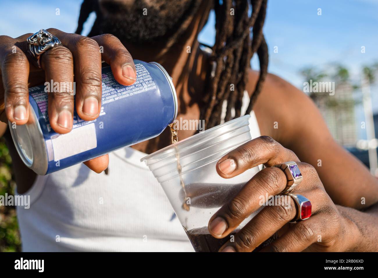 primo piano di una persona di etnia nera irriconoscibile con dreadlock e anelli sulle dita all'aperto al tramonto versando birra in una tazza di plastica, spazio per copiare. Foto Stock