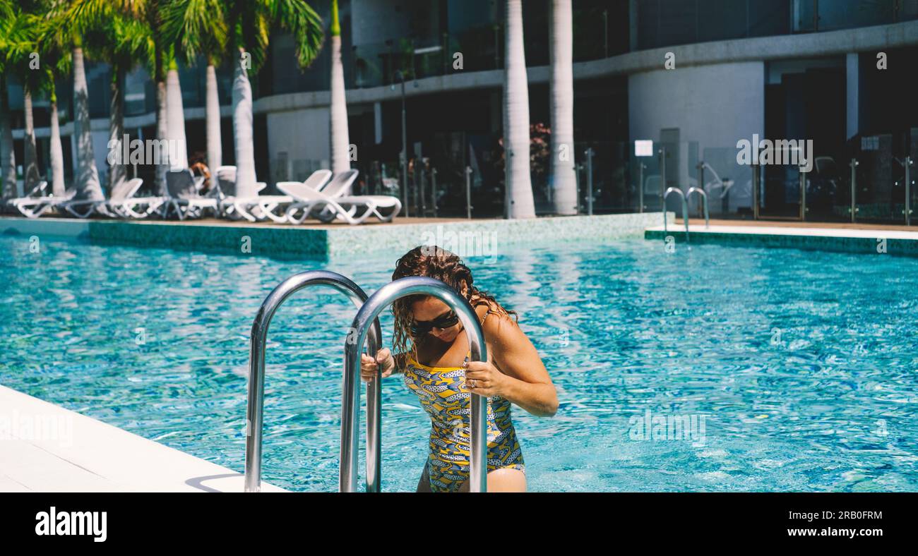 Una ricca signora che esce da una grande piscina blu dopo aver nuotato o essersi rilassata durante le attività estive. Hotel di villeggiatura sullo sfondo. Persone di lusso. Concetto di camere per viaggi e prenotazioni. Banner Foto Stock