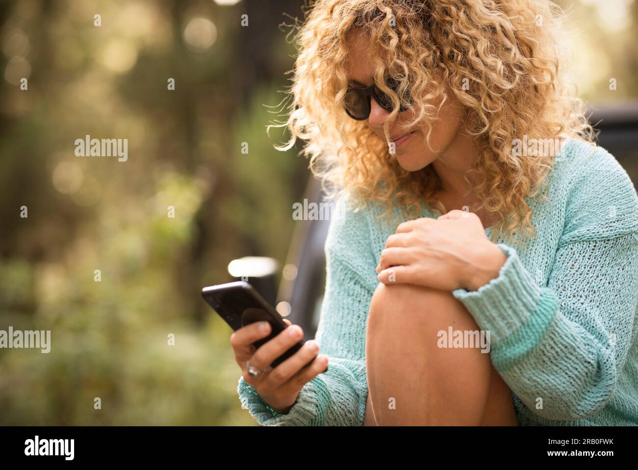 Una bella donna moderna che usa lo scorrimento all'aperto del telefono cellulare e scrive su app. Attività ricreative all'aperto. Giovani donne con capelli ricci biondi che cercano su cellulare. Signora moderna fuori Foto Stock
