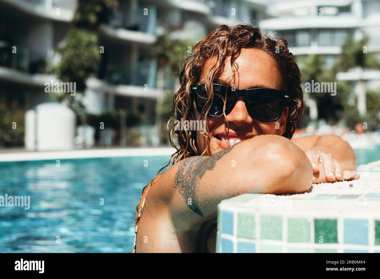 Un turista allegro si goda la piscina e il sole durante le vacanze estive con un resort di lusso in sottofondo. Indossare occhiali da sole. Donne moderne con un sorriso tatuato all'interno dell'acqua della piscina. in salute Foto Stock