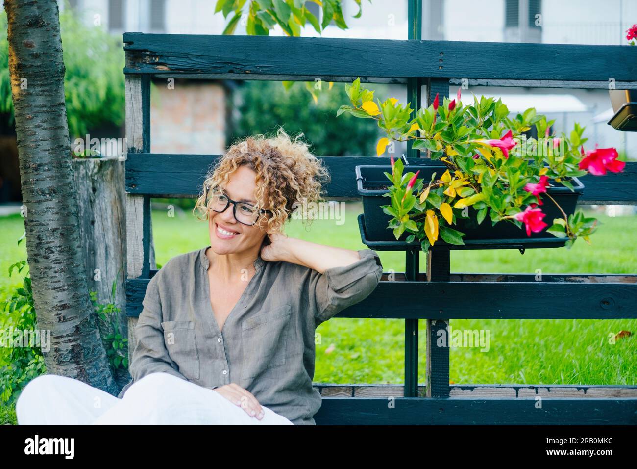 Ritratto di una giovane donna matura che si rilassa e si gode il giardino all'aperto di casa seduto e sorridente. Una donna si rilassa tranquillamente fuori casa. Una felice signora adulta che indossa gli occhiali da vista si siede. Allegro Foto Stock