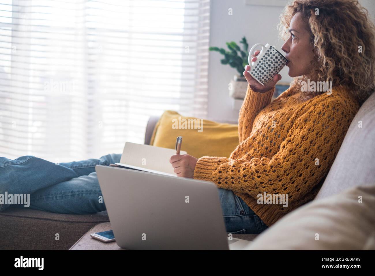Una giovane donna adulta che si rilassa a casa bevendo da una tazza e non scrivendo note con il computer portatile sul divano vicino a lei. Attività ricreative al coperto domenicale. Persone e relax. Una donna che pensa Foto Stock