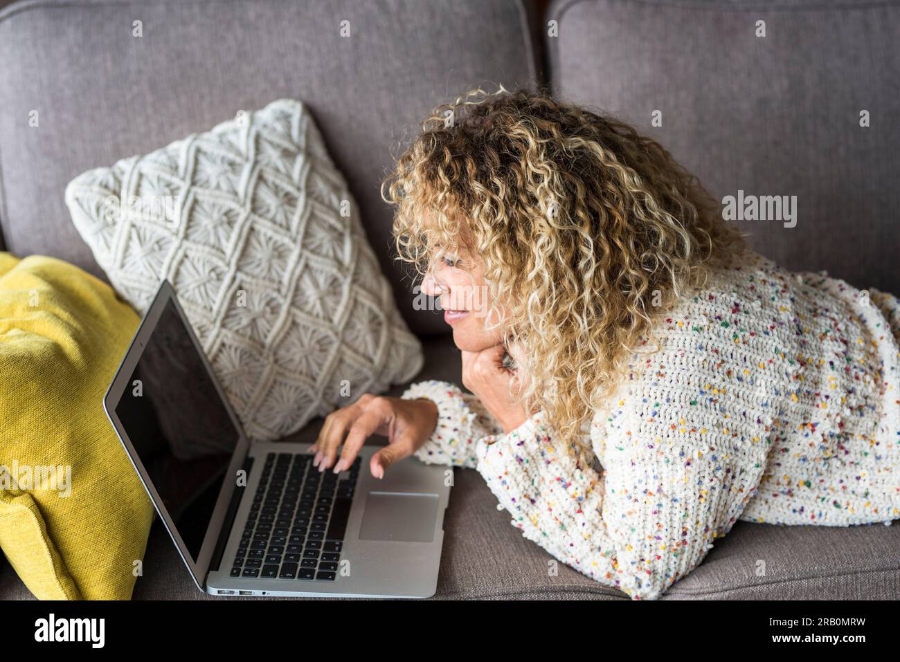 Una giovane donna che utilizza un computer portatile comodamente sdraiata sul divano a casa sorridendo e godendo della tecnologia wireless. Attività ricreative di lavoro in casa. Donne che navigano in rete e si rilassano al coperto Foto Stock