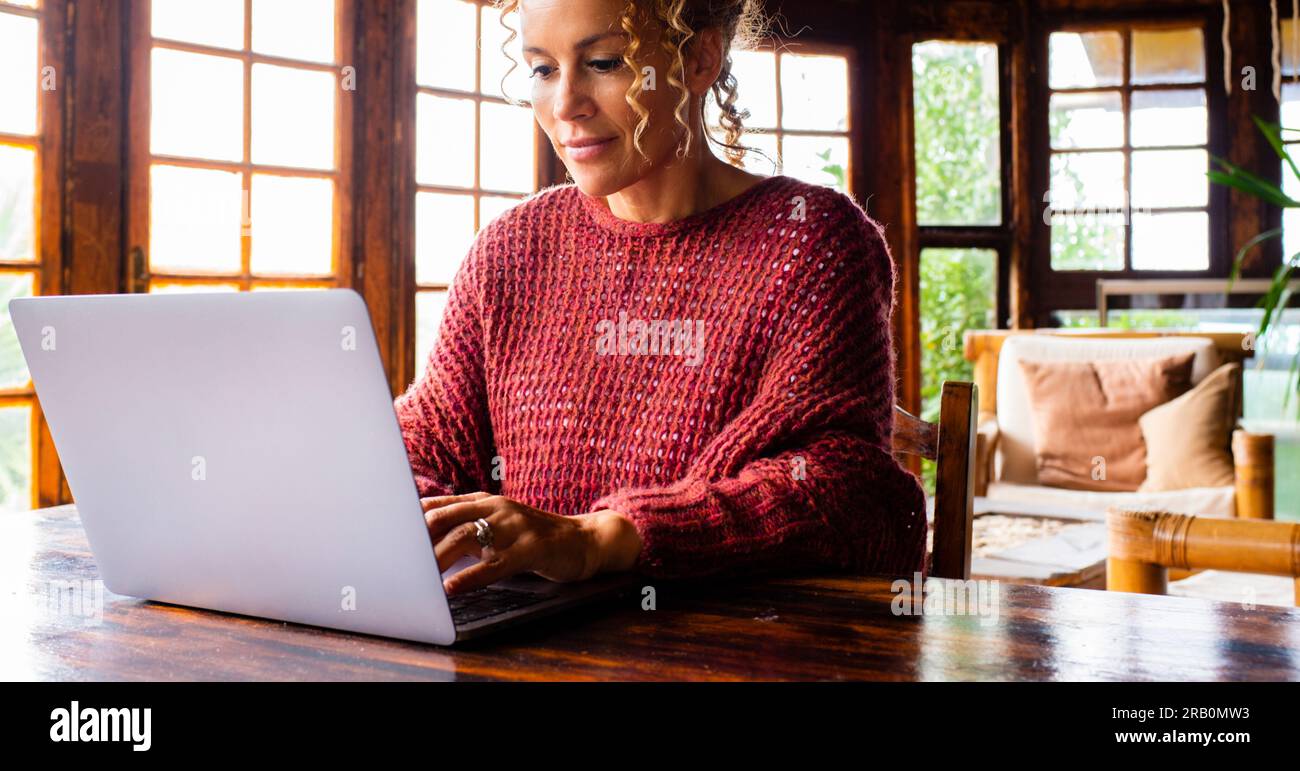 Una donna di mezza età che usa il computer a casa con espressione serena e maglione rosso. Casa interna in legno. Tavolo per il lavoro di ufficio alternativo. Persone che navigano in rete da sole. Viaggi Foto Stock