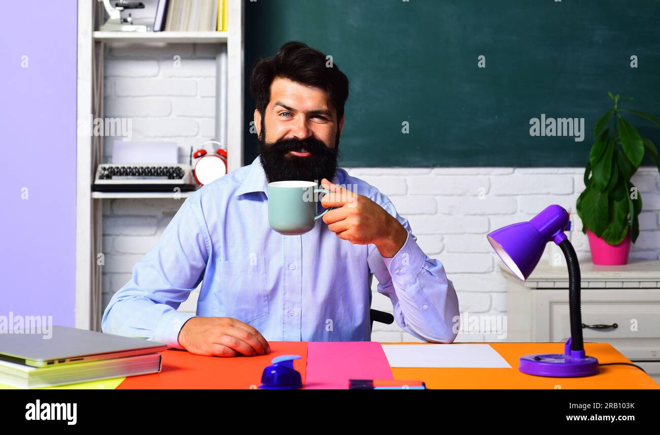 Insegnante maschio sorridente che beve caffè in classe. Insegnante barbuto o professore universitario sul posto di lavoro che beve una tazza di tè. Insegnante di scuola seduta Foto Stock