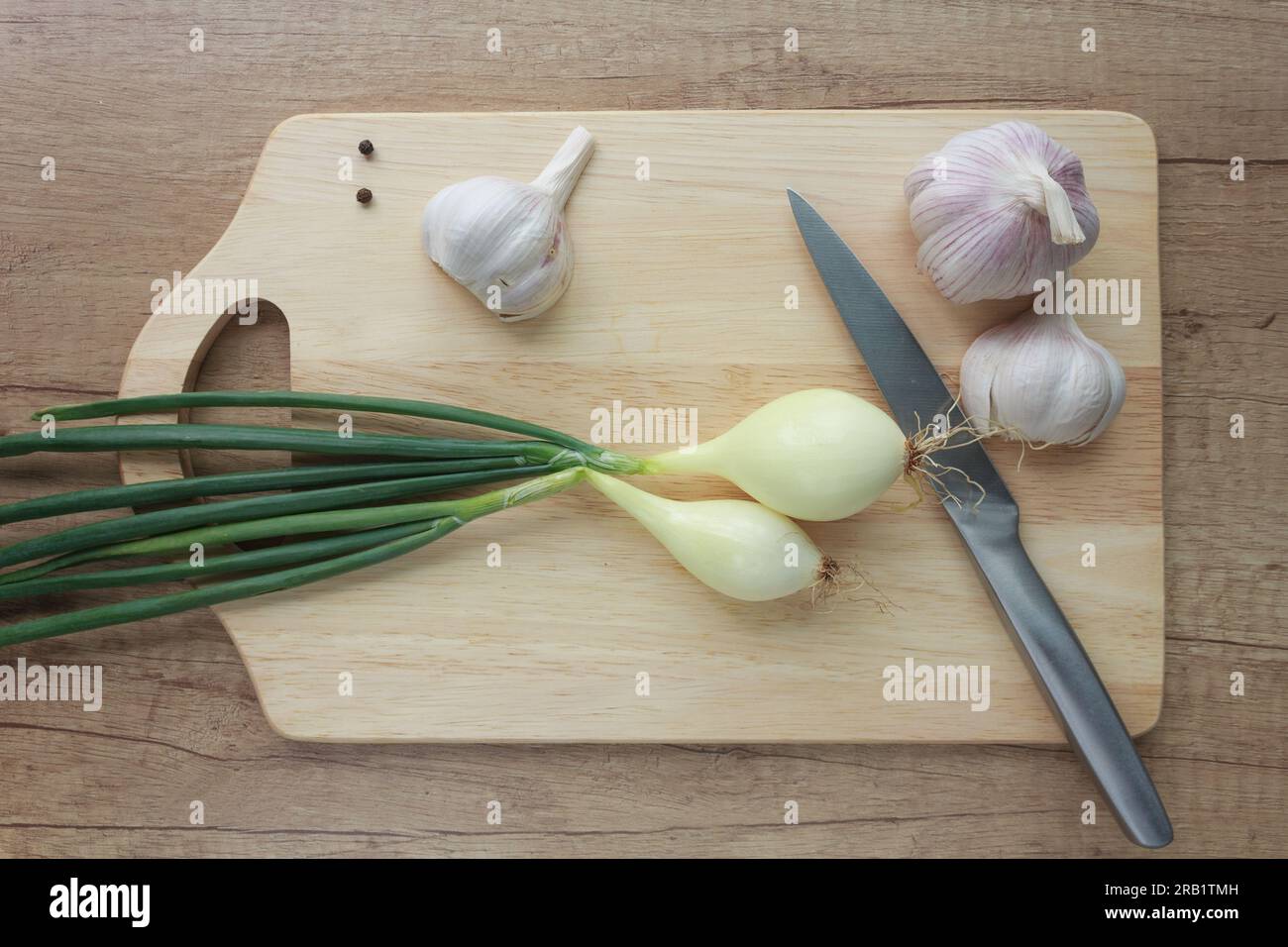 Vista dall'alto di una cipolla giovane pronta per affettare, aglio e grani di pepe nero su uno sfondo di legno. Foto Stock