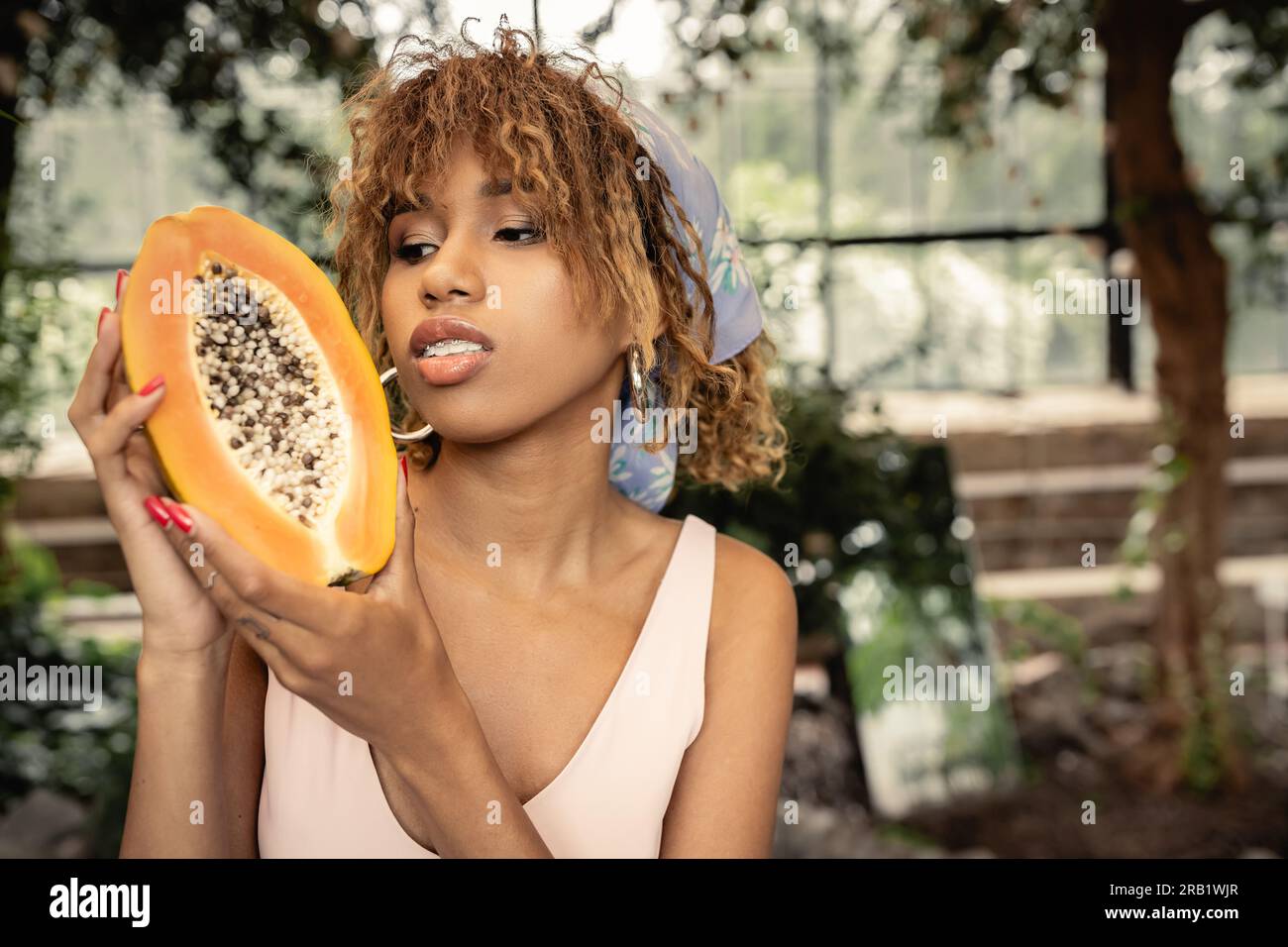 Ritratto di una giovane donna afroamericana alla moda con un velo che regge papaya fresca mentre trascorre del tempo nel centro sfocato del giardino, elegante donna blen Foto Stock