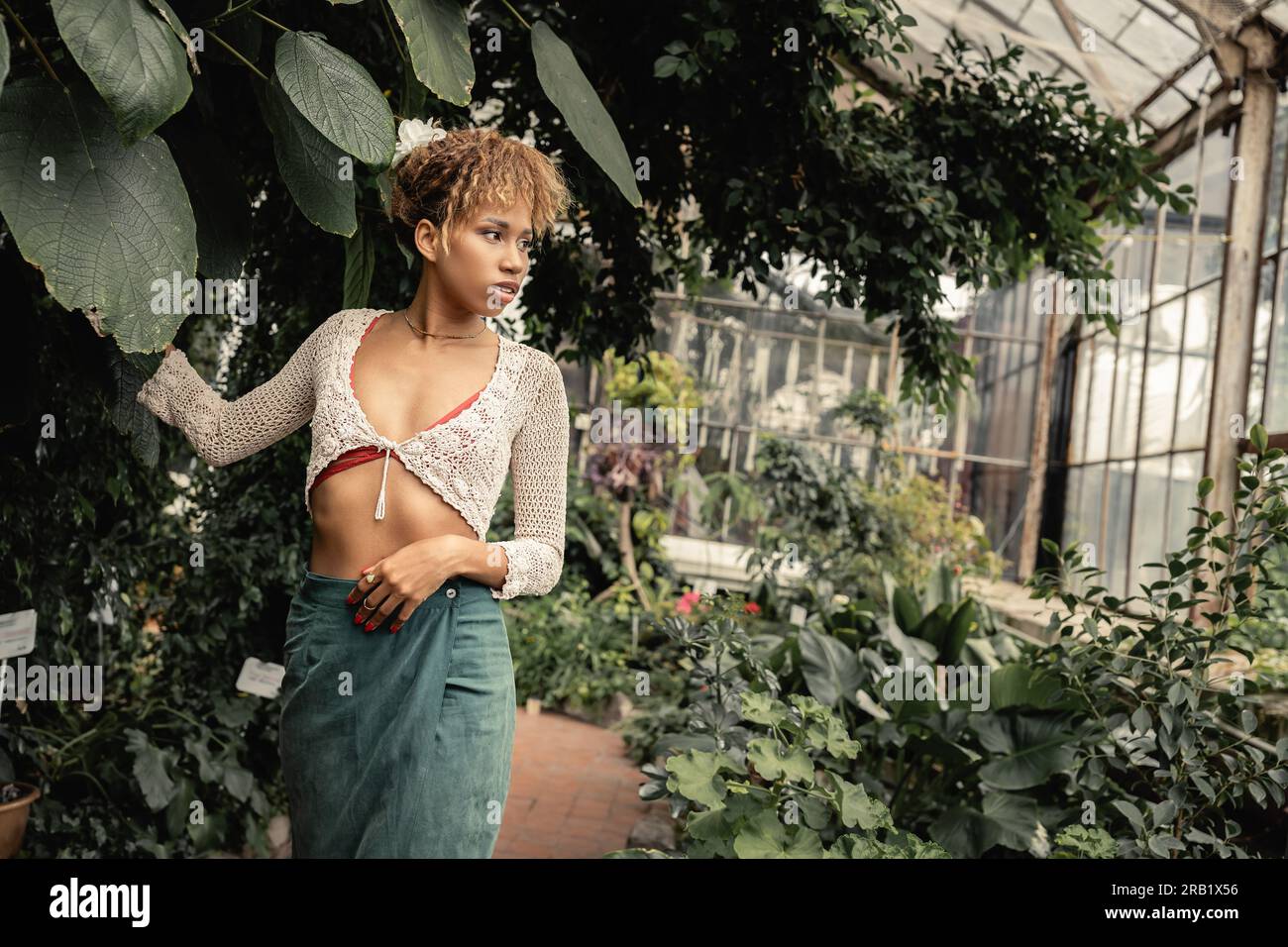 Giovane donna afro-americana sicura di sé con top e gonna in maglia che guarda lontano mentre si trova vicino a piante e foglie nel centro giardino sfocato, fas estivo Foto Stock