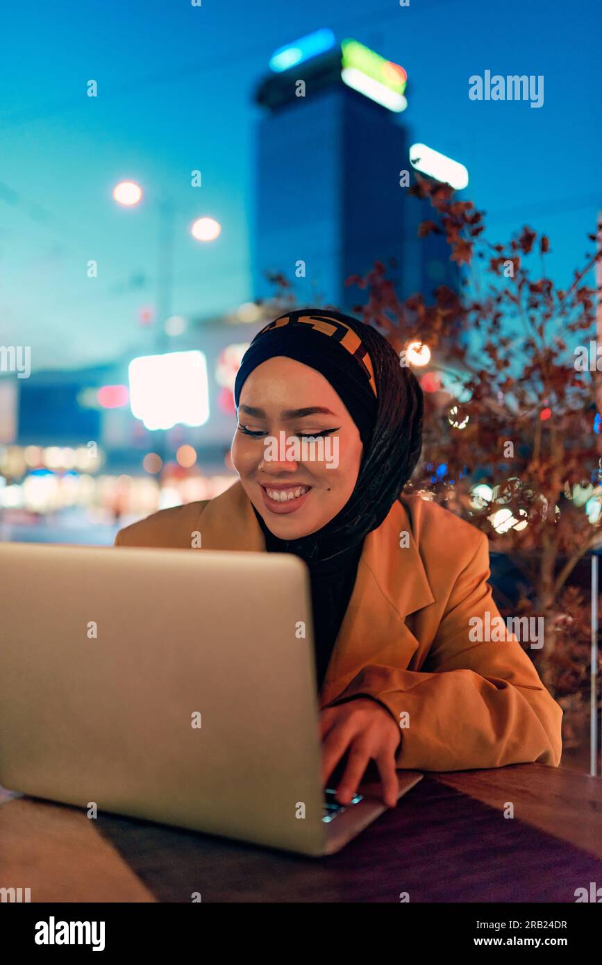 Nell'incantevole atmosfera di un paesaggio urbano notturno, una ragazza rivestita di hijab, coinvolta nel suo laptop, crea un'immagine affascinante, incarnando la fusione di Foto Stock