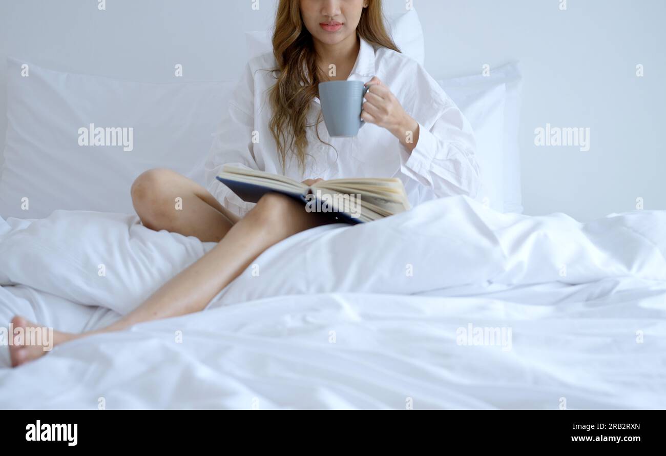 Giovane donna che beve caffè mentre legge un libro seduto sul letto la mattina. Foto Stock