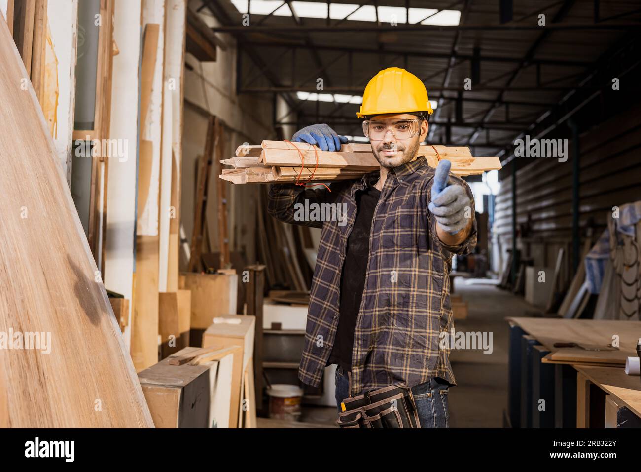 Il lavoro di un operaio edile intelligente trasporta pile di legno in una fabbrica di mobili con i pollici in piedi sorridenti. Foto Stock